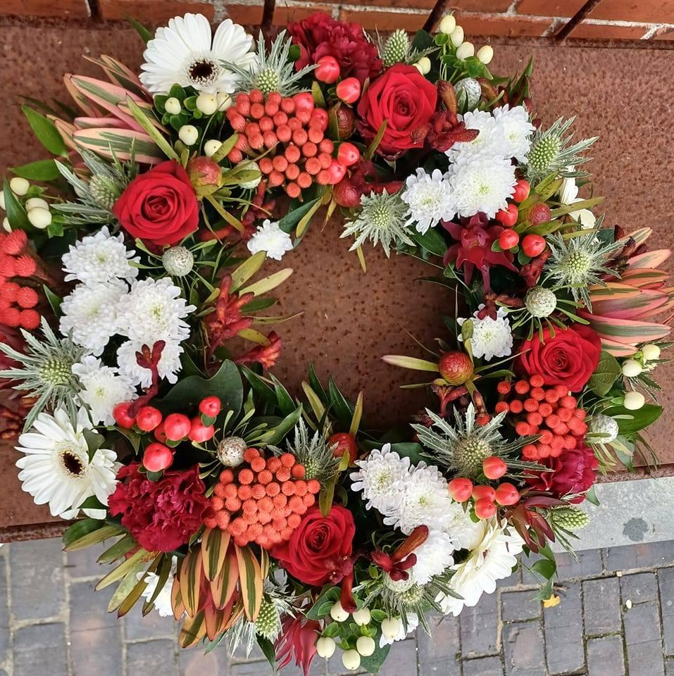 En farverig krans med røde roser, hvide margueritter og forskellige grønne planter, dekoreret med bær og blomster.