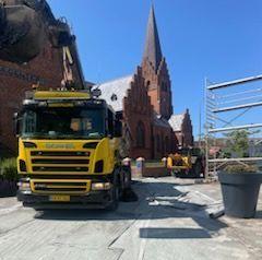 Gul lastbil parkeret foran en kirke under renovering med blå himmel og byggeplads i baggrunden.
