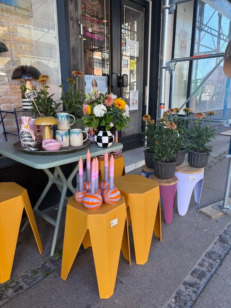Farverige taburetter og en bordopstilling med blomster, stearinlys og dekorative genstande foran en butik.