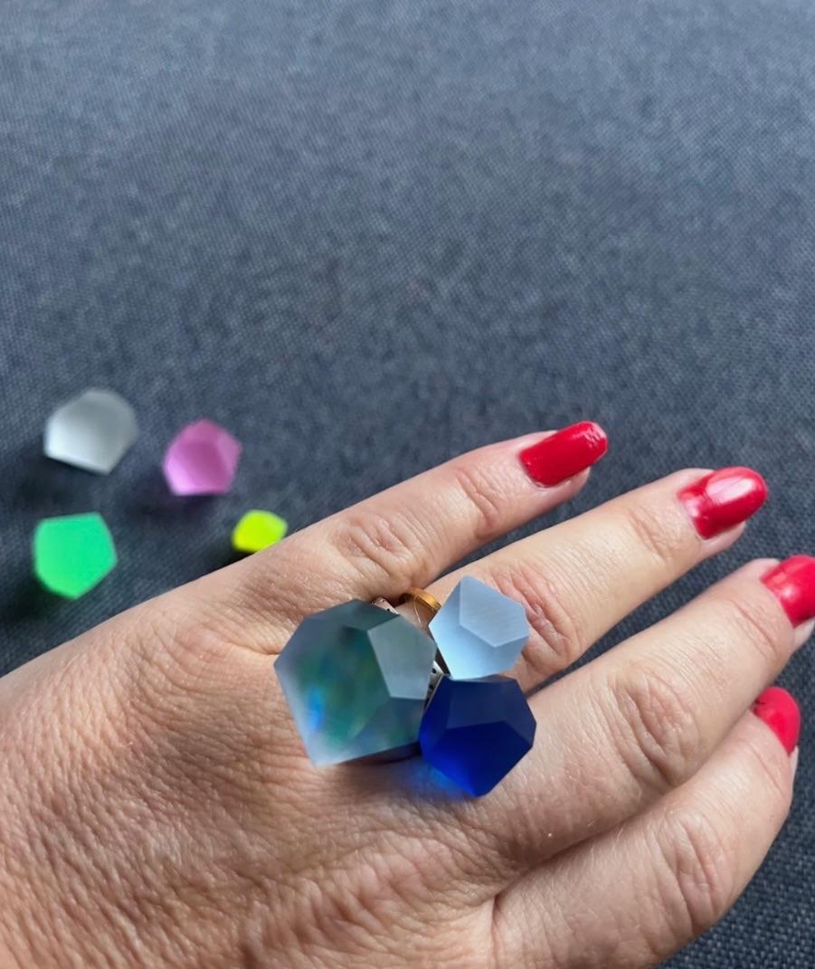 Farverig ring med flere geometriske former i forskellige nuancer, båret på en hånd med røde negle.