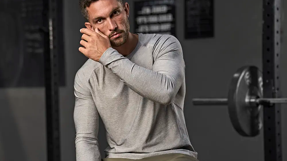 Mand i grå langærmet trøje sidder på en bænk i et fitnesscenter og justerer sit hår.
