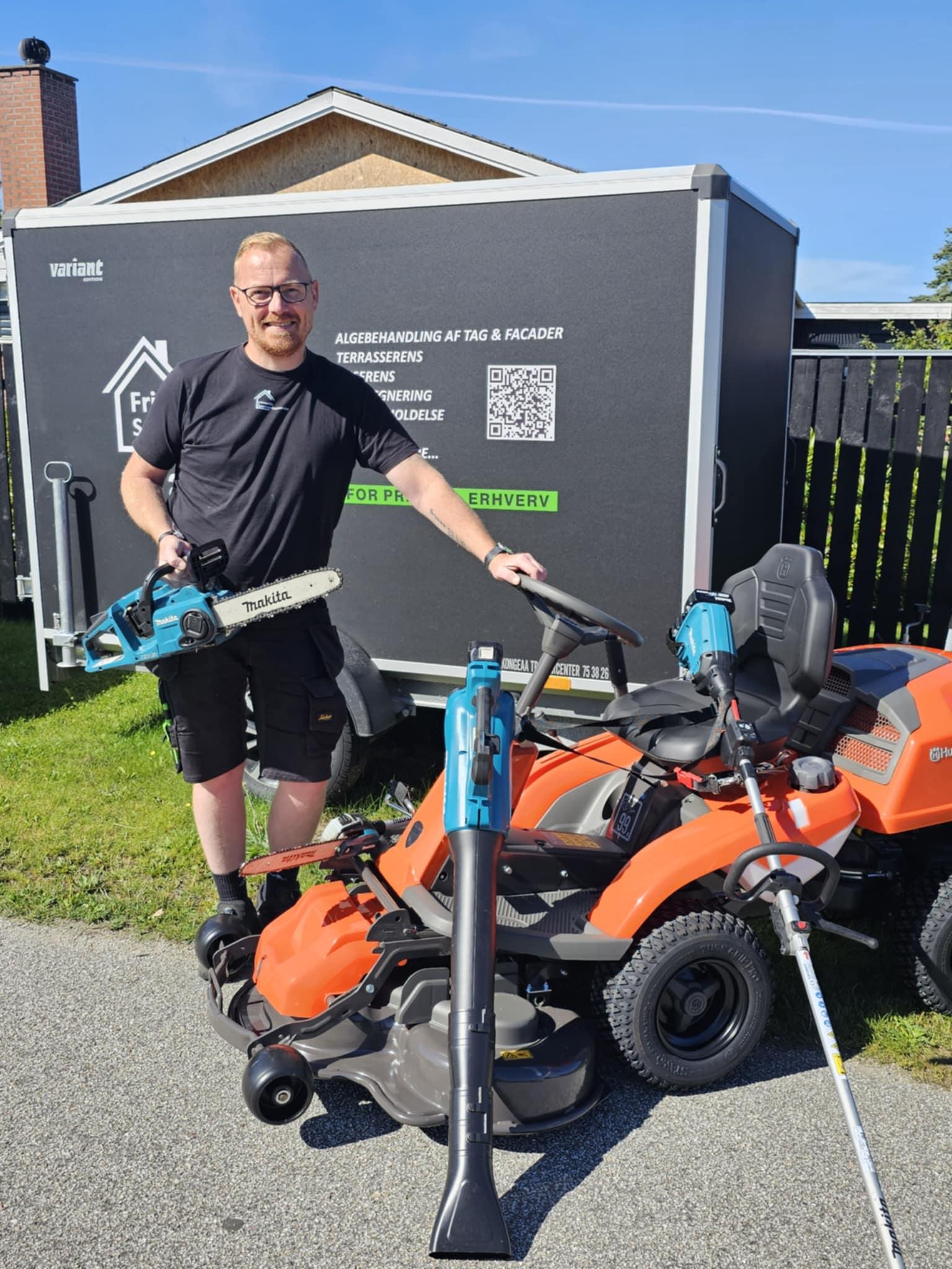 Mand står ved en plæneklipper og en blæser, iført sort t-shirt, med en trailer i baggrunden.