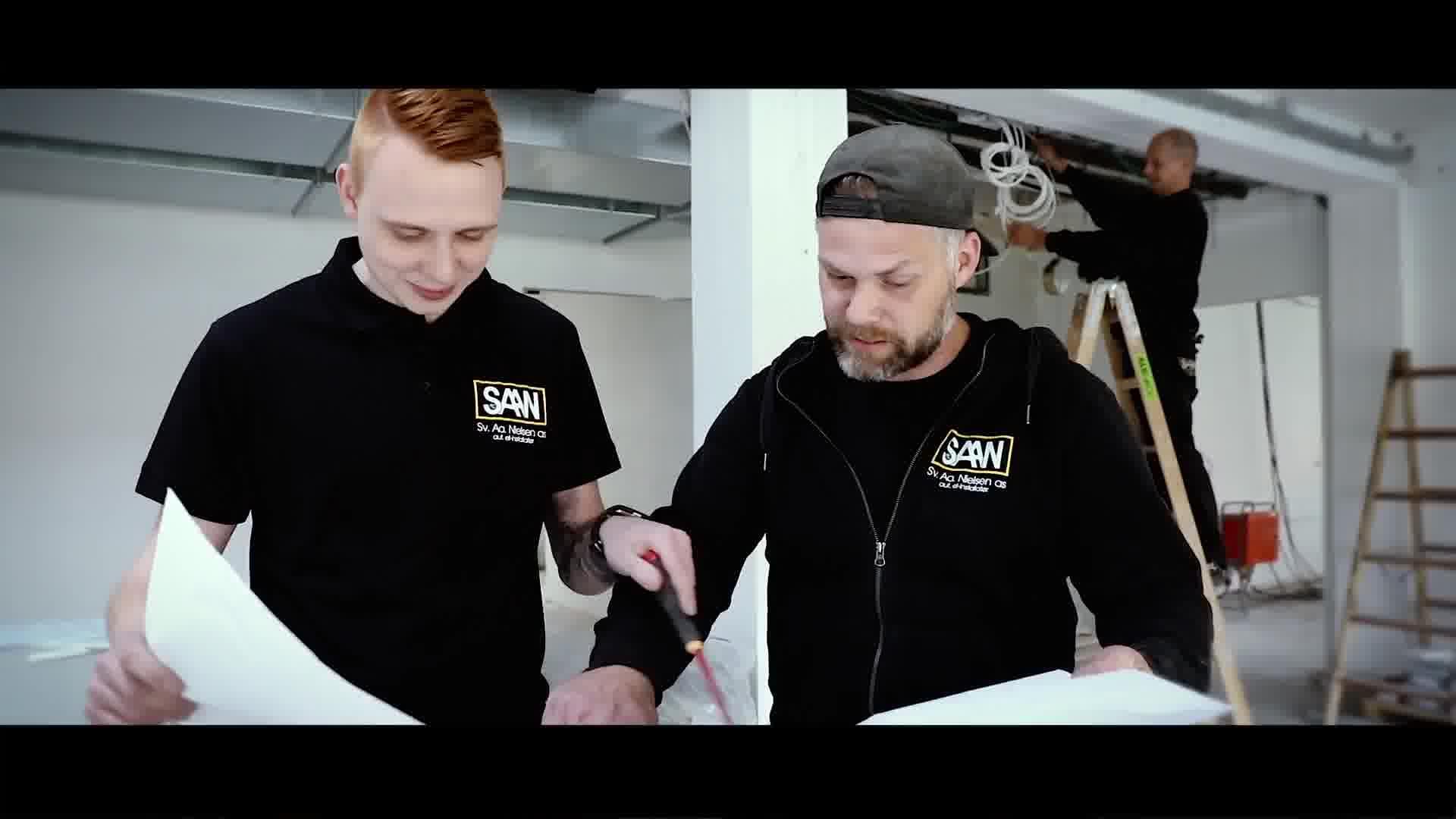 To mænd i sorte T-shirts, der arbejder sammen med malerudstyr i et lyst rum. En mand maler i baggrunden.