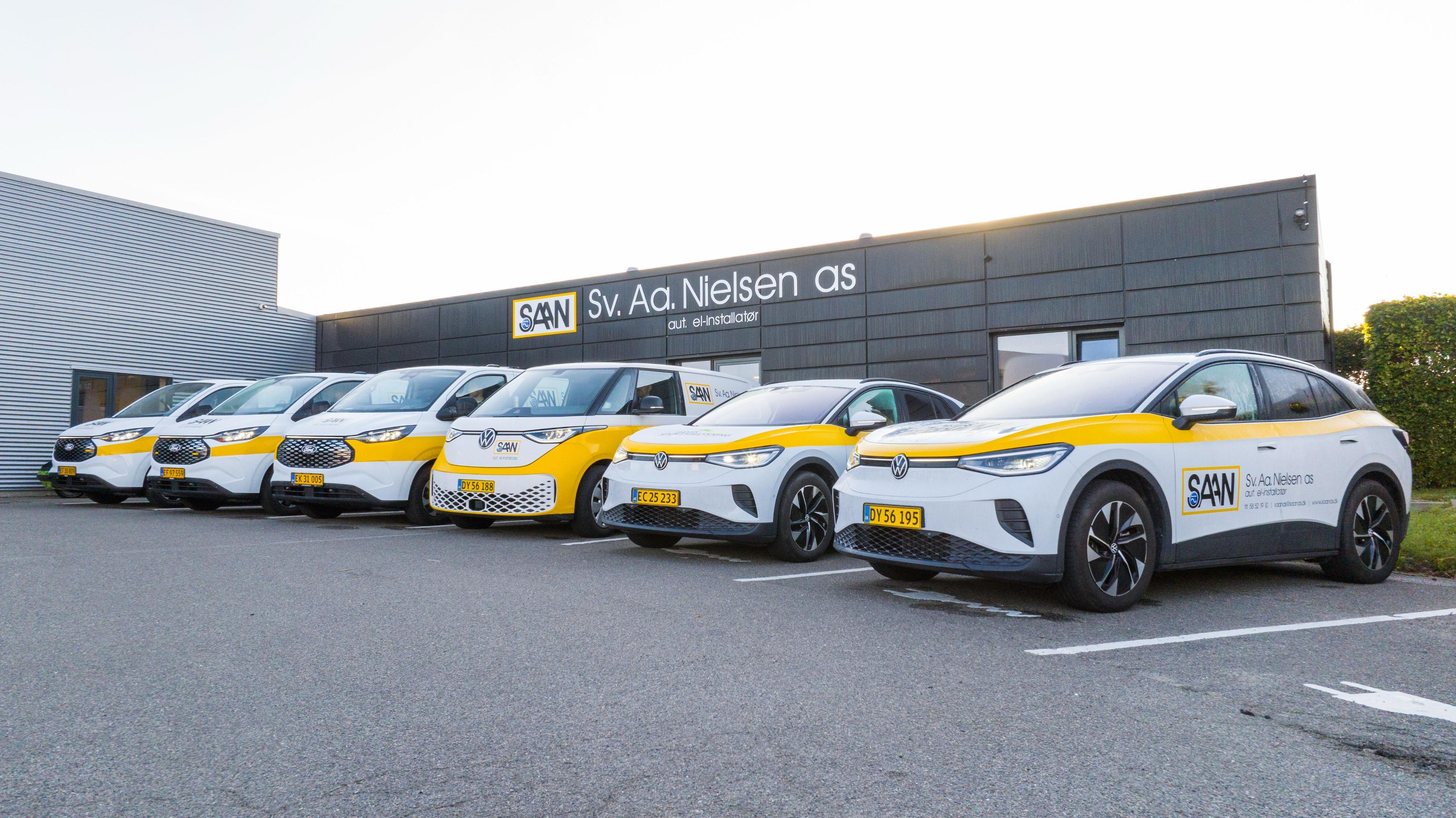 Flere elbiler med logo fra et firma parkeret foran en moderne bygning i dagslys.