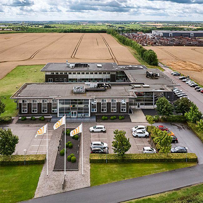 Luftfoto af en moderne kontorbygning med parkeringsplads og marker i baggrunden.