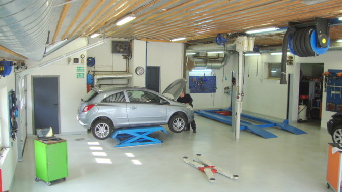 En bilværksted med en sølvfarvet bil på en lift, mekaniker arbejder på motoren, værktøj og udstyr synligt.