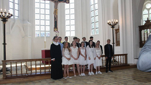 Gruppe af unge mennesker i hvide kjoler og jakkesæt står i en kirke ved en ceremoni med præsten i front.