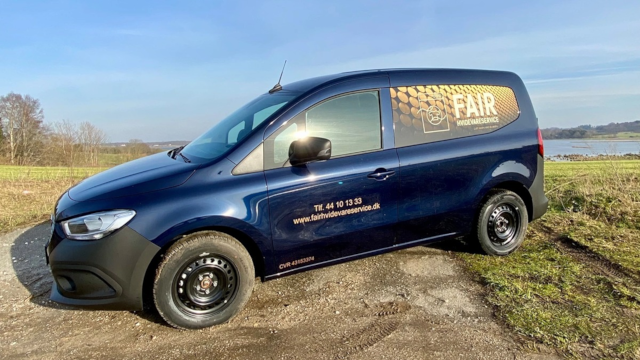 En blå varevogn med reklame for FAIR parkeret ved en grusvej med udsigt over vandet.