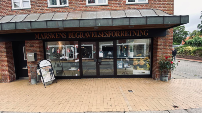 Facade af Marskens Begravelsesforretning med blomster og skilte i en bygade.