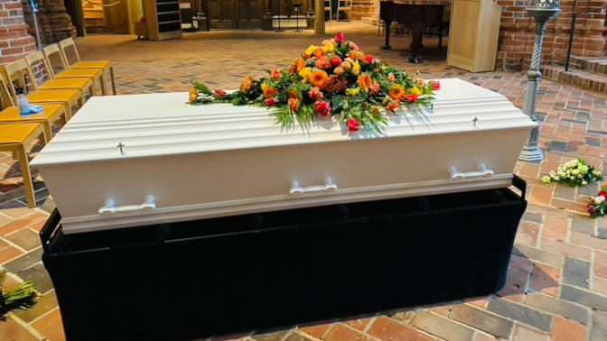 En hvid kiste med farverige blomster på toppen, placeret i en kirke med murstensgulv.