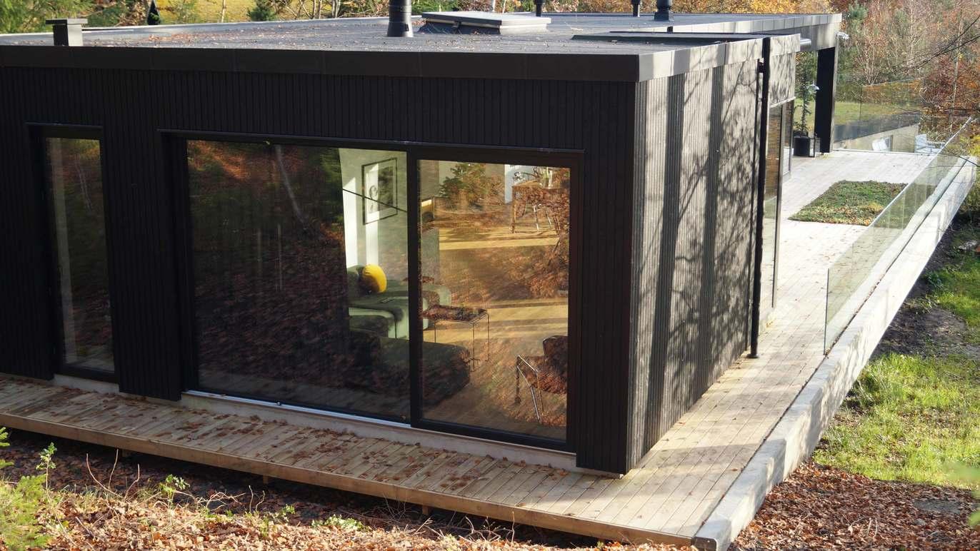 Moderne sort hus med store vinduer og træterrasse omgivet af natur.