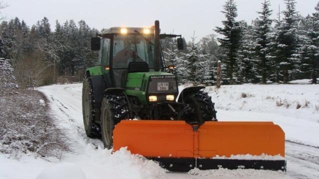 En traktor med en orange sneplov rydder sne på en vej i en skovklædt vinterlandskab.