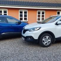To biler parkeret ved en bygning, en blå SUV og en hvid SUV, med grus under dem.