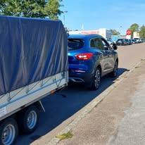 En blå bil trækker en trailer med presenning på en parkeringsplads under en klar himmel.