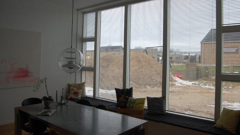 Moderne stue med store vinduer, der viser en byggeplads og en spisebord med stole og puder.