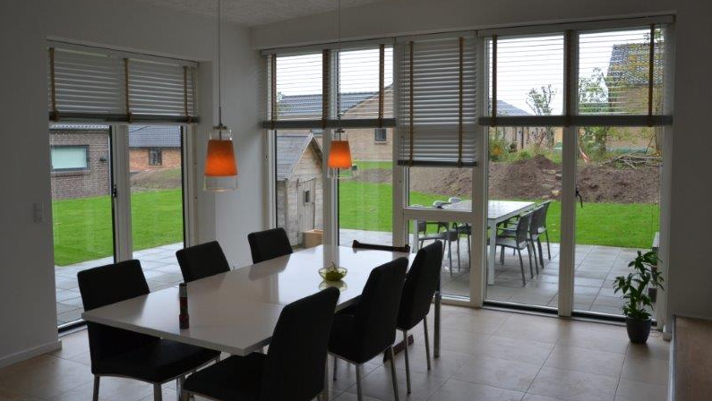 Moderne spisestue med stort bord, sorte stole og udsigt til haven gennem store vinduer.