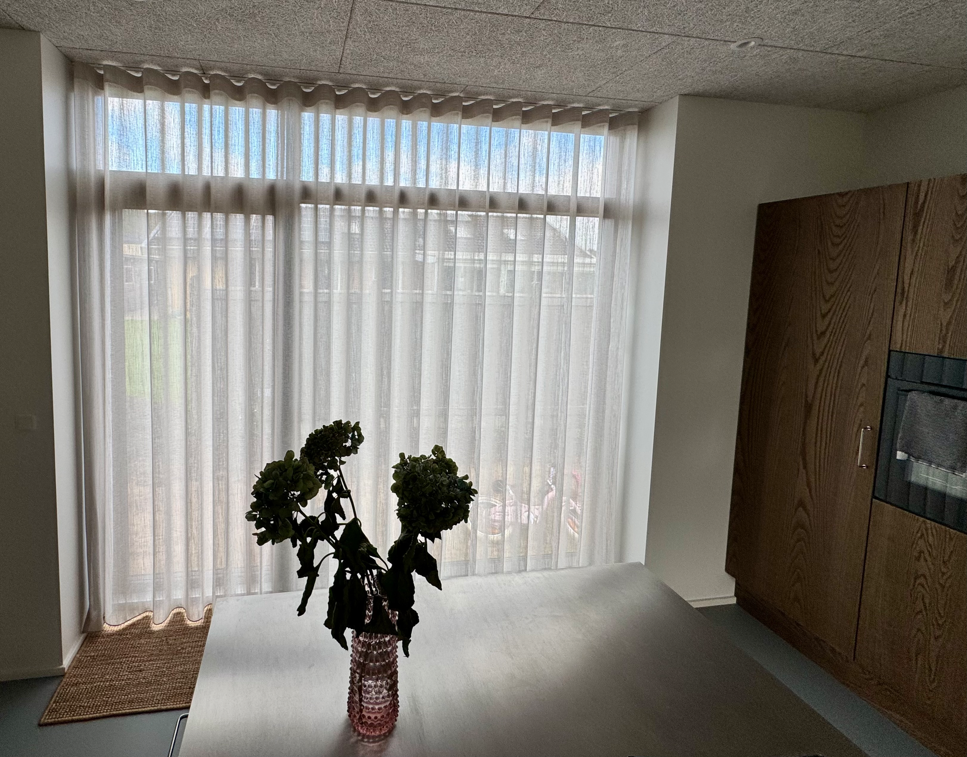 Lys og luftig stue med store vinduer, hvide gardiner og en vase med blomster på et bord.