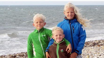 Tre børn står på stranden med havet i baggrunden, iført farverige jakker og smilende ansigtstræk.