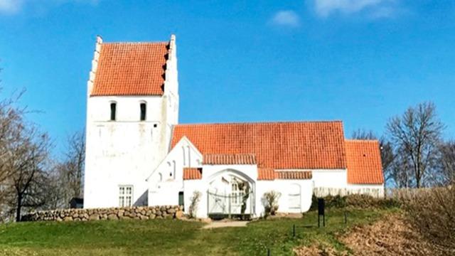 En hvid kirke med rød tagsten beliggende på en bakke under en blå himmel.