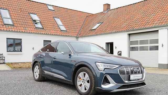 En blå Audi E-Tron SUV parkeret foran et hus med rødt tag og vinduer i taget.