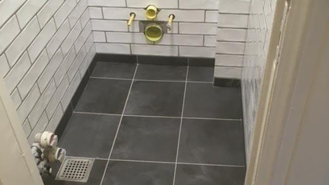 Badeværelse med grå fliser, hvidt murværk og synlige rør til vandinstallationer.