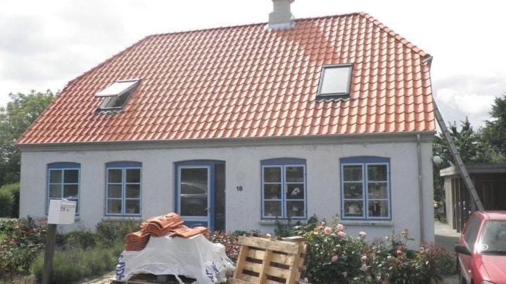 Et hvidt hus med rødt tag, blå vinduer og en gård fyldt med materialer og blomster.