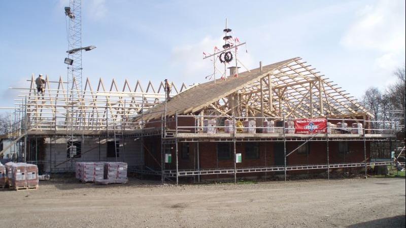 Byggeri af et hus med tagkonstruktion, stillads og arbejdere på stedet under en klar himmel.