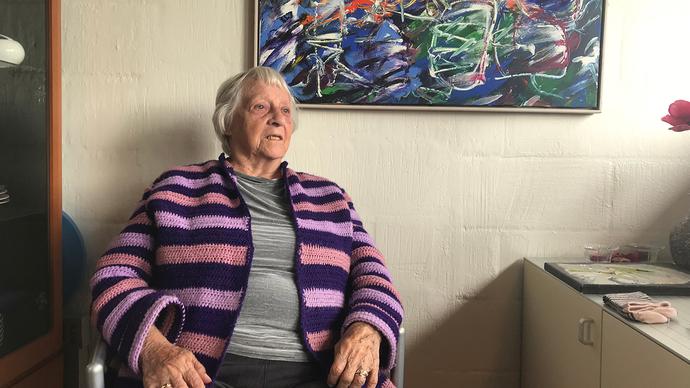 Ældre kvinde i stribet sweater sidder i en stol foran et farverigt maleri i stuen.