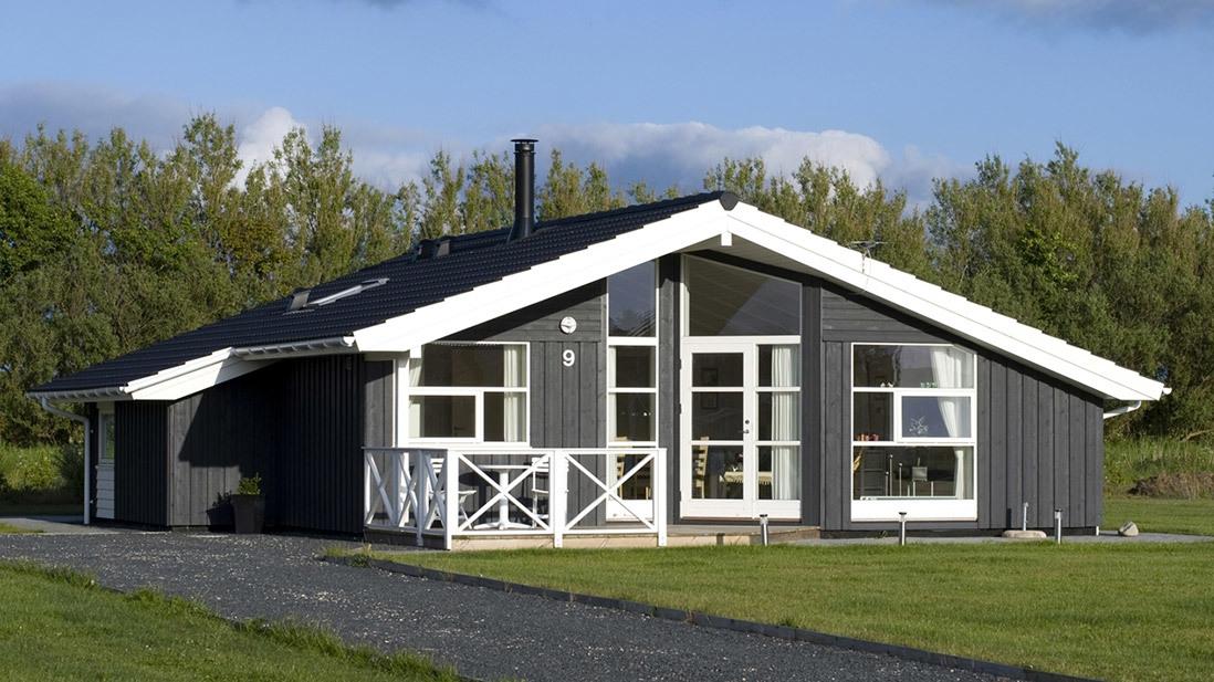 Moderne sommerhus med sort facade og hvidt tag, beliggende i et grønt område med græsplæne.