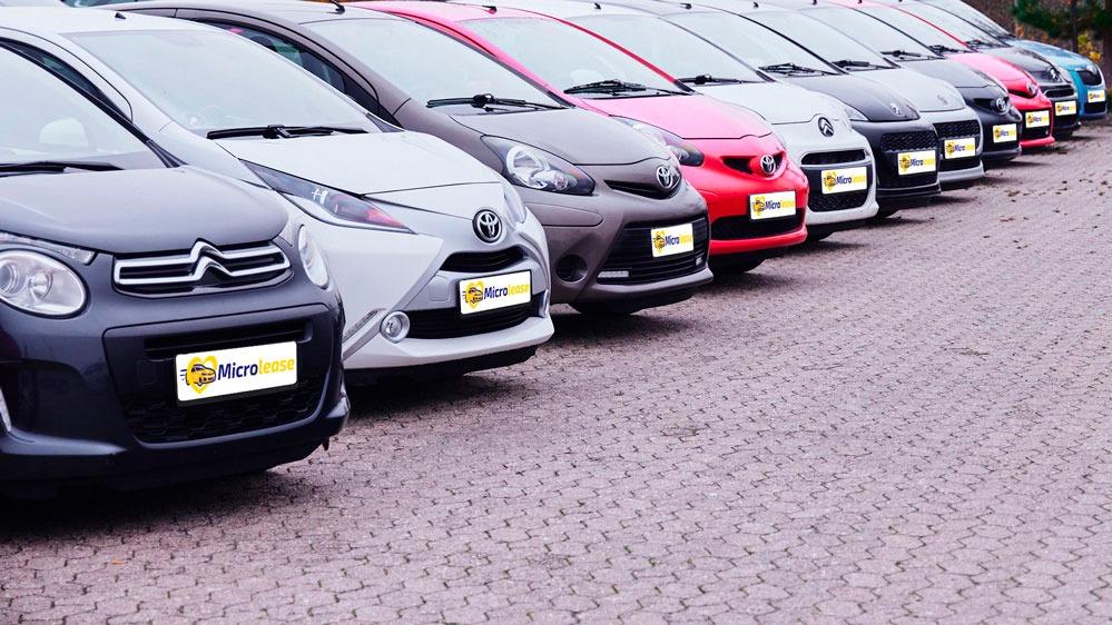 Række af farverige biler parkeret på en brostensbelagt plads, herunder grå, hvid, rød og sort.