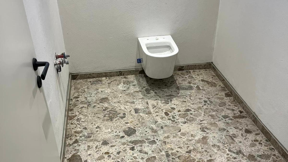 Badeværelse med en hvid toilet og grå fliser på gulvet. Vægge i lys farve.