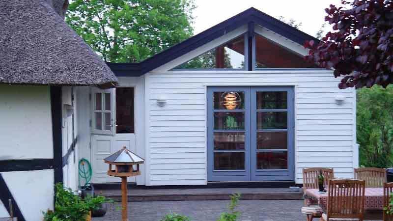 Moderne hvidt hus med skråtag, store vinduer og terrasse med træmøbler i en grøn have.
