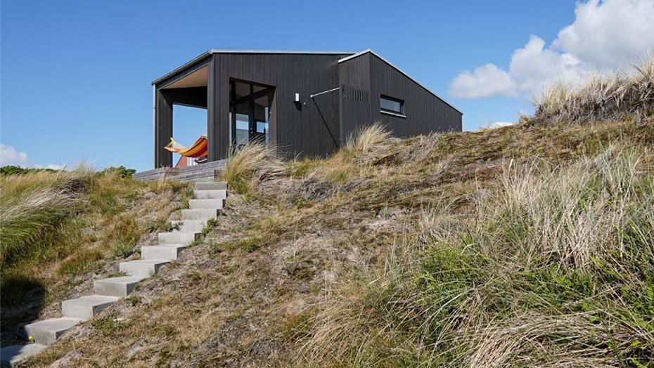 Moderne sommerhus på en skråning med græs og stentrappe, omgivet af natur og blå himmel.
