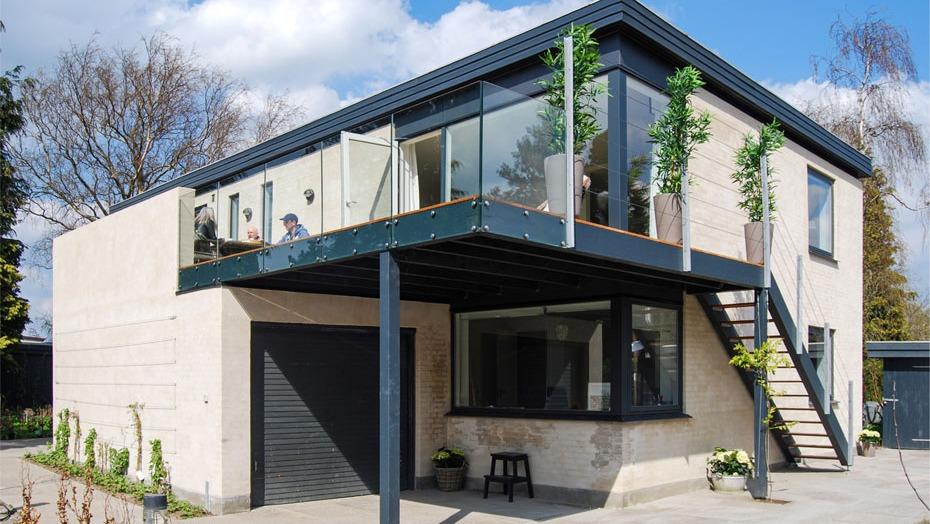 Moderne hus med glasaltan, sort ramme og grønne planter, beliggende i et roligt kvarter.