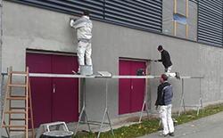 Tre malere arbejder på en væg, mens de står på stilladser, med en rød dør i baggrunden.