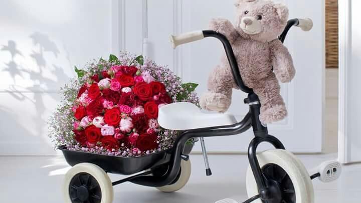 En trehjulet cykel med en bamse og en stor buket røde roser i bagagerummet.