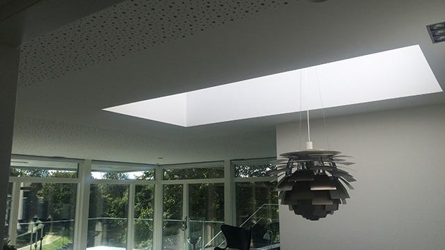 Lysindfald fra ovenlysvindue i moderne stue med en unik lampe og store vinduer.