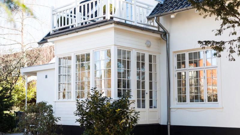 Hvidt hus med store vinduer og en balkon, omgivet af grønne buske og træer.
