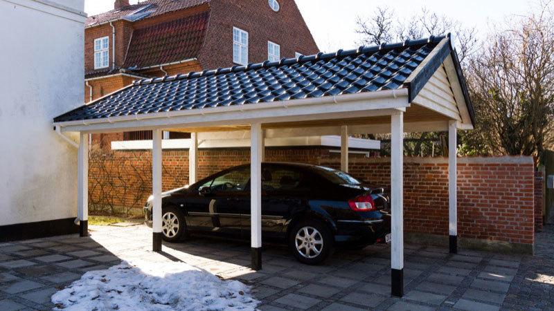 En sort bil parkeret under en carport med tag i fliser og en murstensvæg i baggrunden.