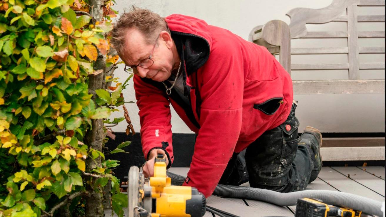 En håndværker i rød jakke arbejder på en terrasse med en elektrisk sav og en støvsuger ved siden af.