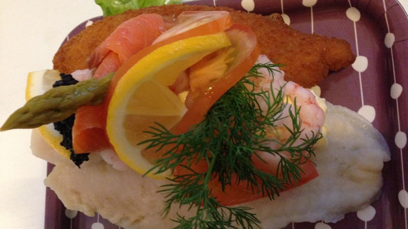 En tallerken med paneret fisk, rejer, tomater, citron, asparges og dild på en brun bakke.
