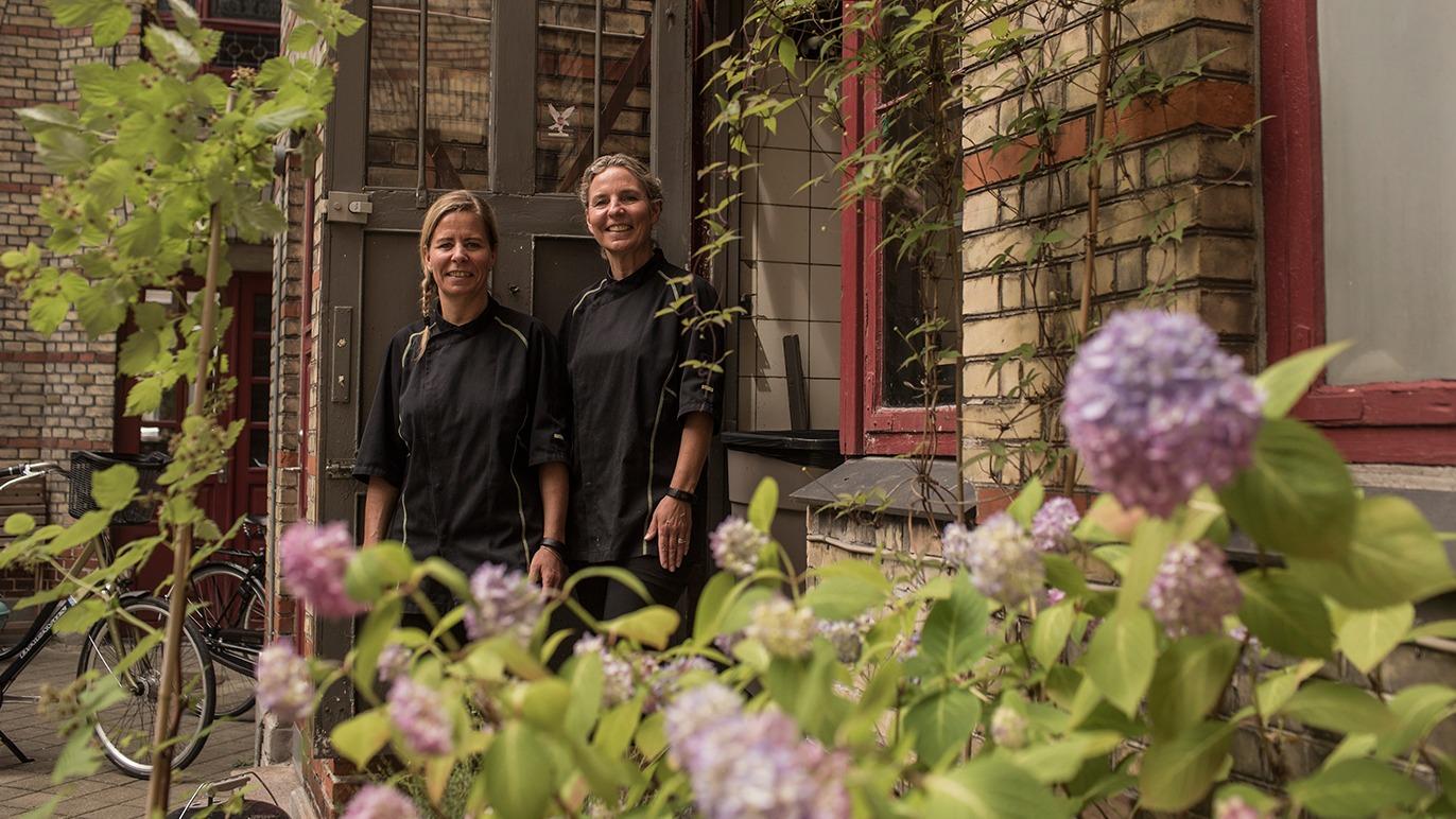To personer i sorte kokkejakker står foran en murstensvæg med blomster i forgrunden.