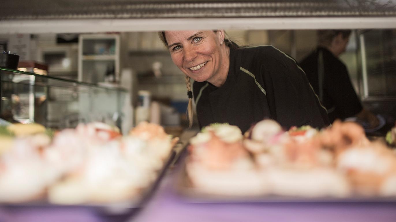En smilende kvinde står bag en disk med farverige sushi-retter i en restaurant.