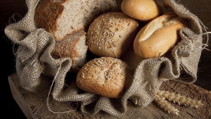 Friskbagt brød og rundstykker i en jutepose på en træplade med hvedeaks i baggrunden.