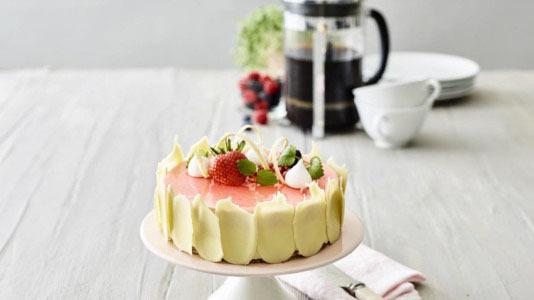 En smuk lagkage med jordbær og flødeskum, serveret med kaffe og tallerkener i baggrunden.