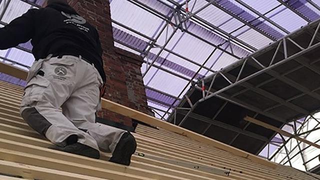Håndværker arbejder på et tag med træplanker under en bygning med et beskyttende tag.