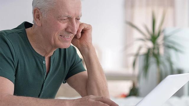 En glad ældre mand sidder foran en bærbar computer og smiler, mens han arbejder hjemme.