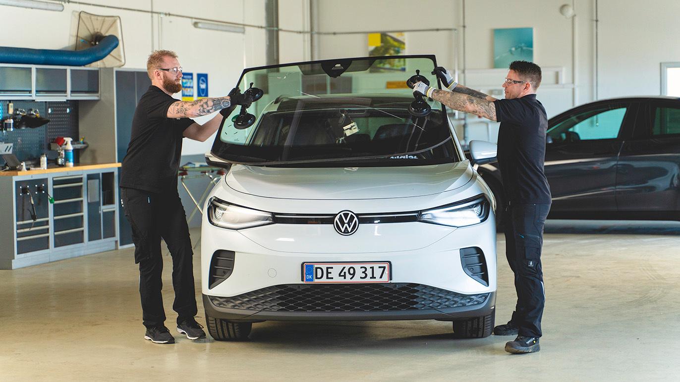 To personer, der arbejder sammen om at installere forruden på en hvid Volkswagen ID.4 i et værksted.