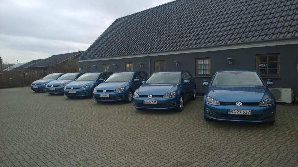 Seks blå Volkswagen biler parkeret foran en grå bygning med tag af sorte teglsten.