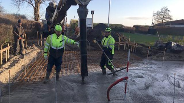 To arbejdere i sikkerhedstøj arbejder på en byggeplads med beton og jernstænger. En gravemaskine er i baggrunden.